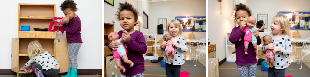 Two children stand holding baby dolls in the dramatic play area. One child puts away a baby doll. Another child holds a baby doll as she waits to put it away.