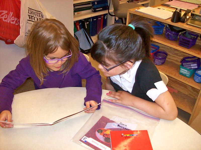Two children sit at a table and read a book together.