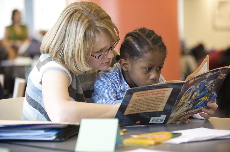 A teacher sits behind a child and points out text features as the child reads a book at a table.