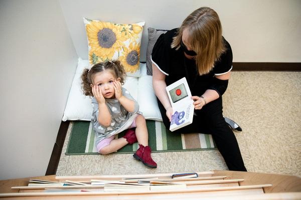 A teacher sits on a pillow on the floor and shows a child sitting on a mat pictures in a cardboard book.