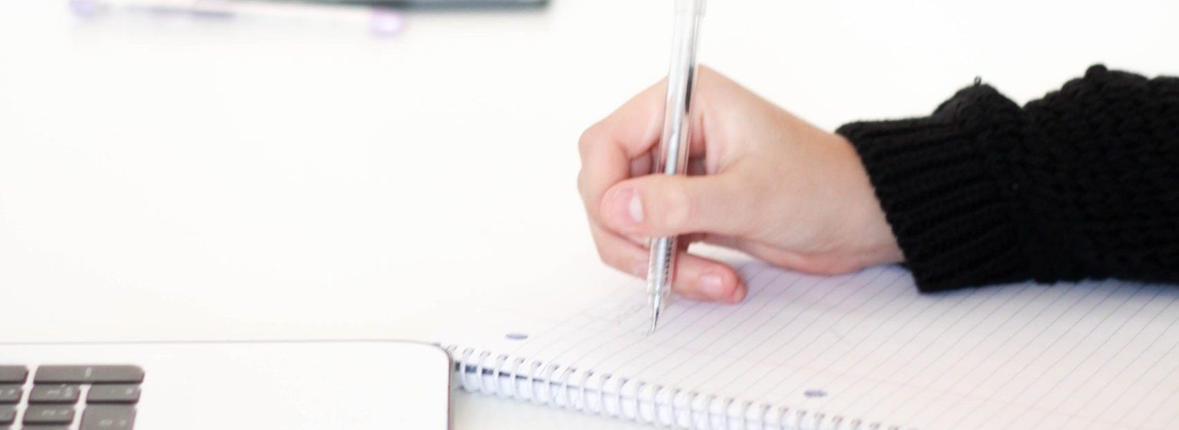 photo showing part of a laptop keyboard and a notebook. There is a hand holding a pen writing on the notebook. In the background there is a sign saying Life is a Journey Enjoy IT