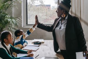 Teacher and two students around a table with papers on it. Teacher is standing and giving a high five to one of the students.