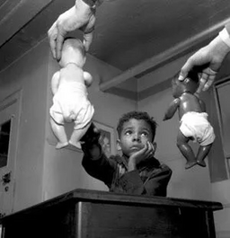 In the foreground is the hands of a researcher, each holding a baby doll in the air in front of a black boy, about age 8. One doll has light skin and the other has dark skin.