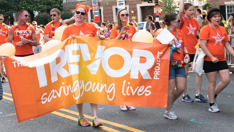 Several people walking in a parade, all wearing orange shirts with stars on them. A large banner is held by several people, it reads "The Trevor Project, Saving Young Lives"