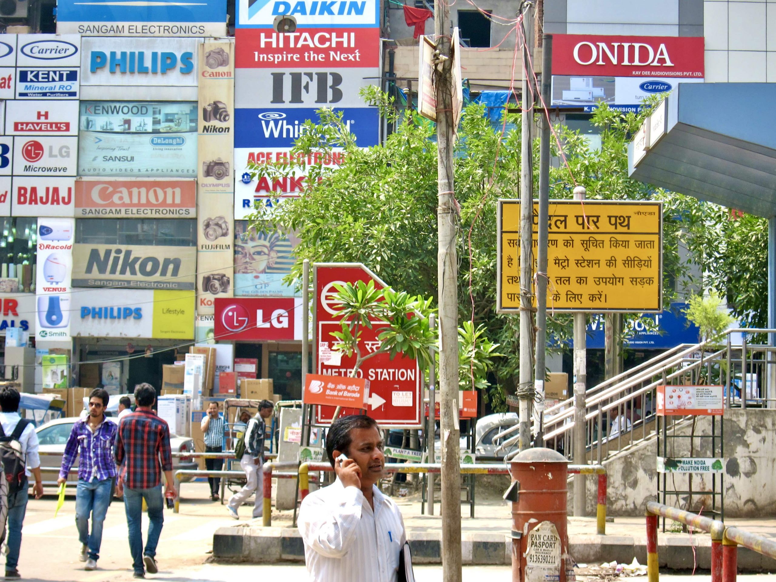 Man in foreground on mobile phone on a busy street in India, with backdrop of signs with Japanese, Korean, and US logos and brands brands like LG, Nikon, Canon, Whirlpool, and Hitachi