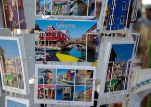 A photograph of a postcard rack, with seven different types of postcards on it. Each postcard shows different scenery. The main postcard in focus in the middle of the picture. It shows a red building to the left with a bridge over water to another building to the right, with a yellow boat in the foreground.
