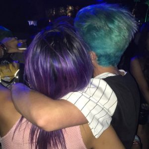 A photograph of two people standing beside each other, viewed from behind. The person on the left has long, purple colored hair. The person on the right has short, light blue colored hair, and their arm is around the person on the left.
