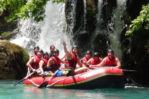 A photograph of nine people in helmets and lifejackets, sitting in a raft in the water. The person in front has their hand raised in a wave toward the camera. In the background is a waterfall and greenery.