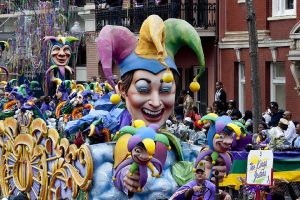 A photograph of a parade during Mardi Gras in New Orleans. The photograph is centered on a parade float shaped like a jester holding two smaller jesters. All three jesters have purple, yellow, and green hats. More identical jesters are in the background, along with a crowd of people and brick buildings.
