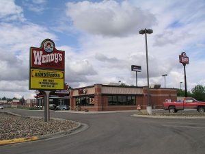 A photograph of a Wendy’s Restaurant, rectangular building with a flat roof, viewed from the road. Pole mounted Wendy’s sign in front.