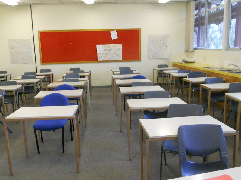 A classroom has isolated desks in straight rows with a mostly-empty bulletin board at the back.
