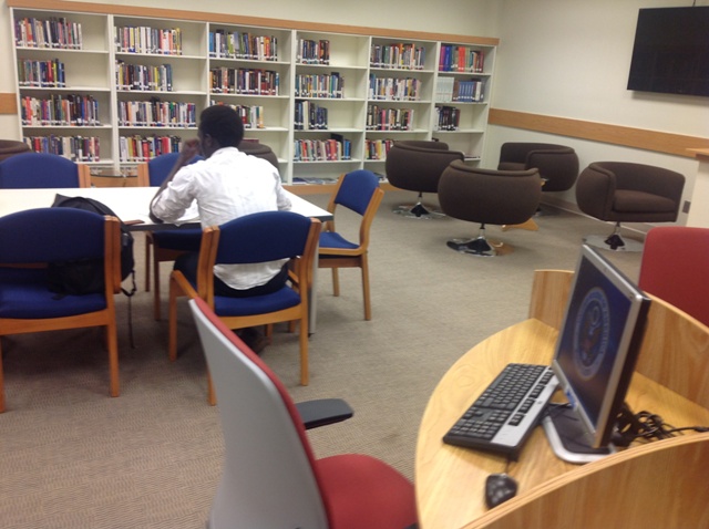 A room has a bookshelf with books at the back. In front of the bookshelf, a student sits at a table with five chairs. The room also has four rolling chairs and a computer workstation.